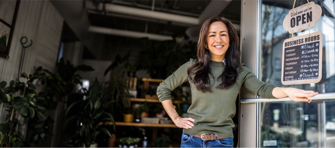 A female small business owner smiling and holding open the glass door to her shop.