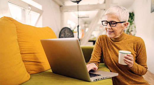 An older woman with a coffee mug in one hand, sitting down and looking at a laptop screen.