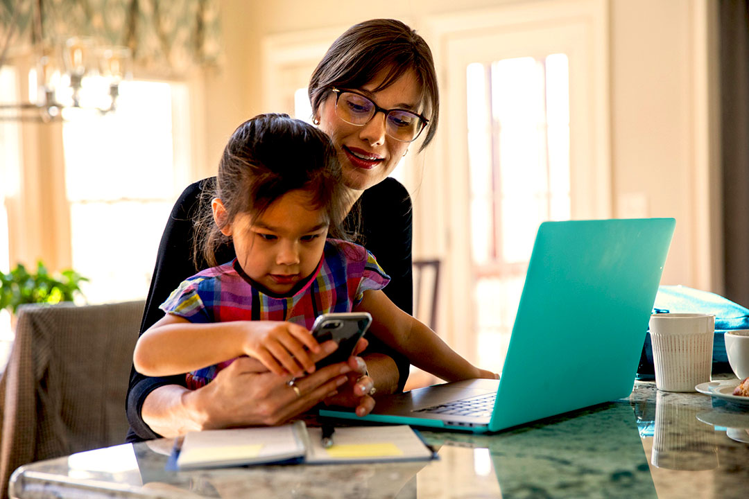 Daughter sitting on a mother's lap playing with a cell phone, while mom works on a laptop.