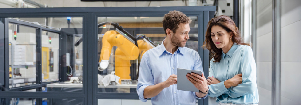 A male and female coworker review a tablet in front of a robotics lab.